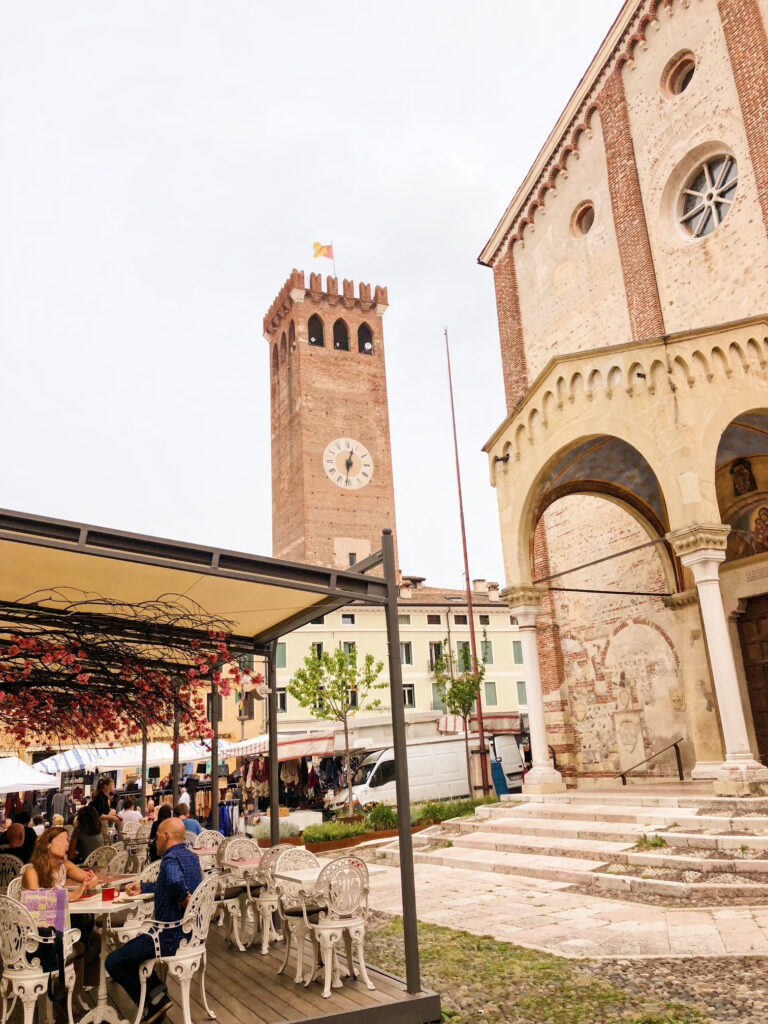 Al fresco diners enjoying lunch beside the Duomo di Santa Maria in Bassano del Grappa, with the town’s medieval clock tower rising in the background.