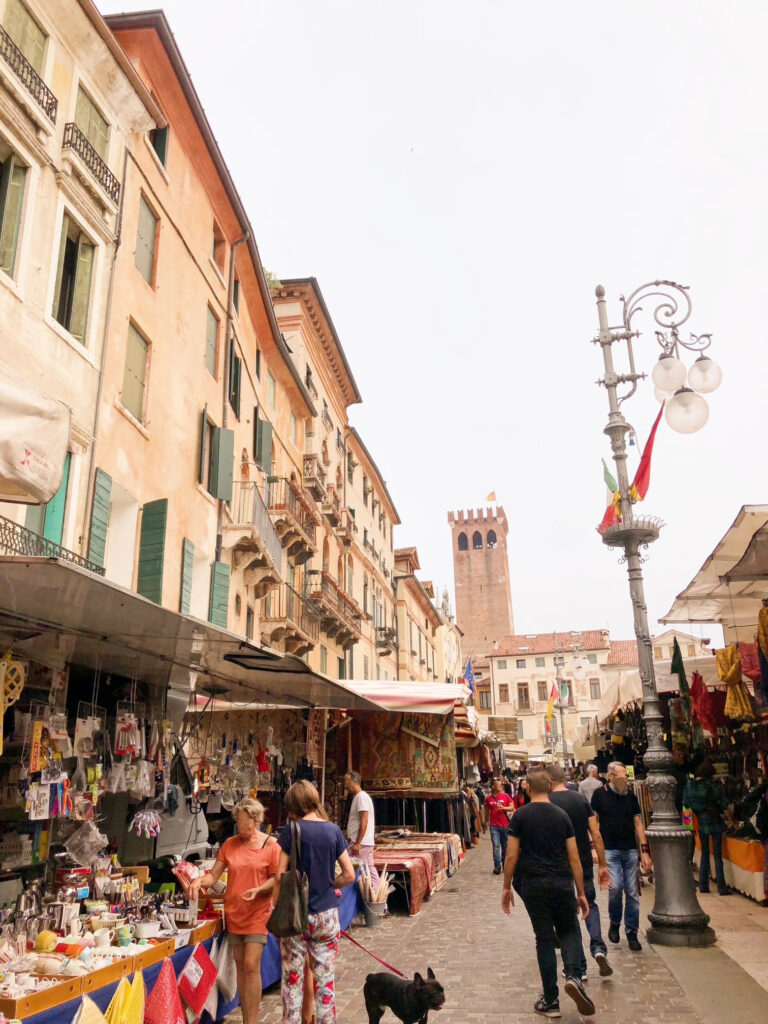 Busy Thursday market in Bassano del Grappa, Italy, with locals browsing colourful textile and antique stalls beneath historic buildings and a medieval tower.