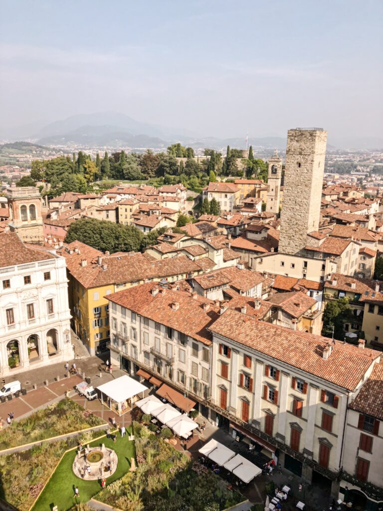 Aerial view of Piazza Vecchia and medieval tower in Città Alta, Bergamo, Italy, with terracotta rooftops and the Alps in the background.