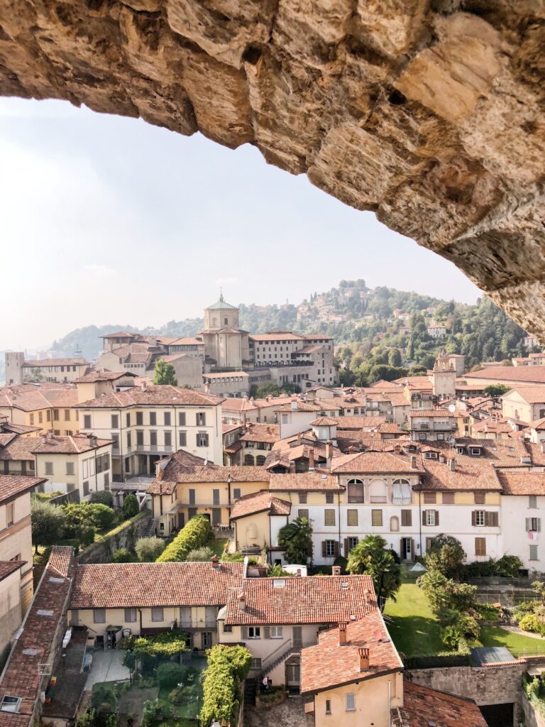 Panoramic view of Bergamo’s Upper Town rooftops framed by a stone arch, with rolling green hills and distant villas in the Lombardy countryside.