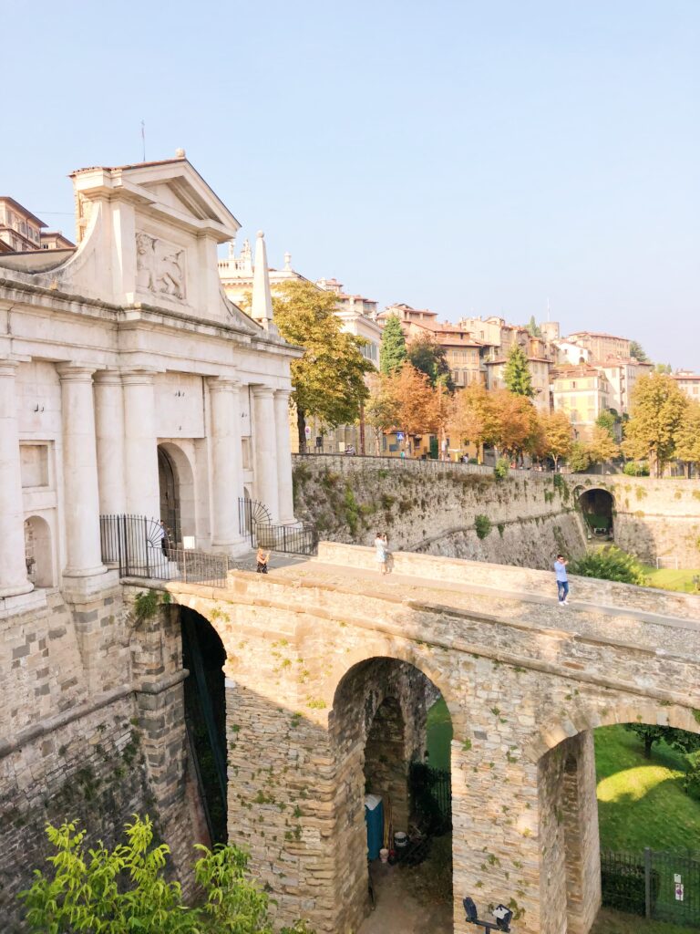 Historic stone gate and Venetian walls of Città Alta in Bergamo, Northern Italy, on a sunny autumn day with visitors strolling across the arched bridge.