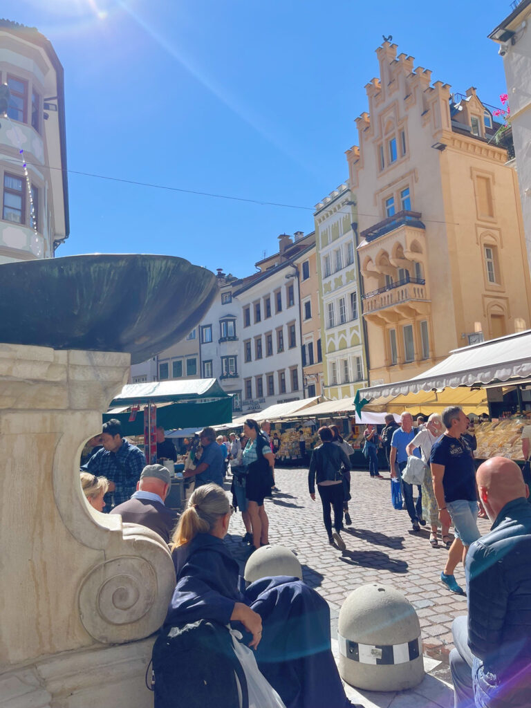 Bustling market scene in Bolzano’s historic centre with people shopping beneath striped awnings, surrounded by ornate buildings and alpine sunshine.