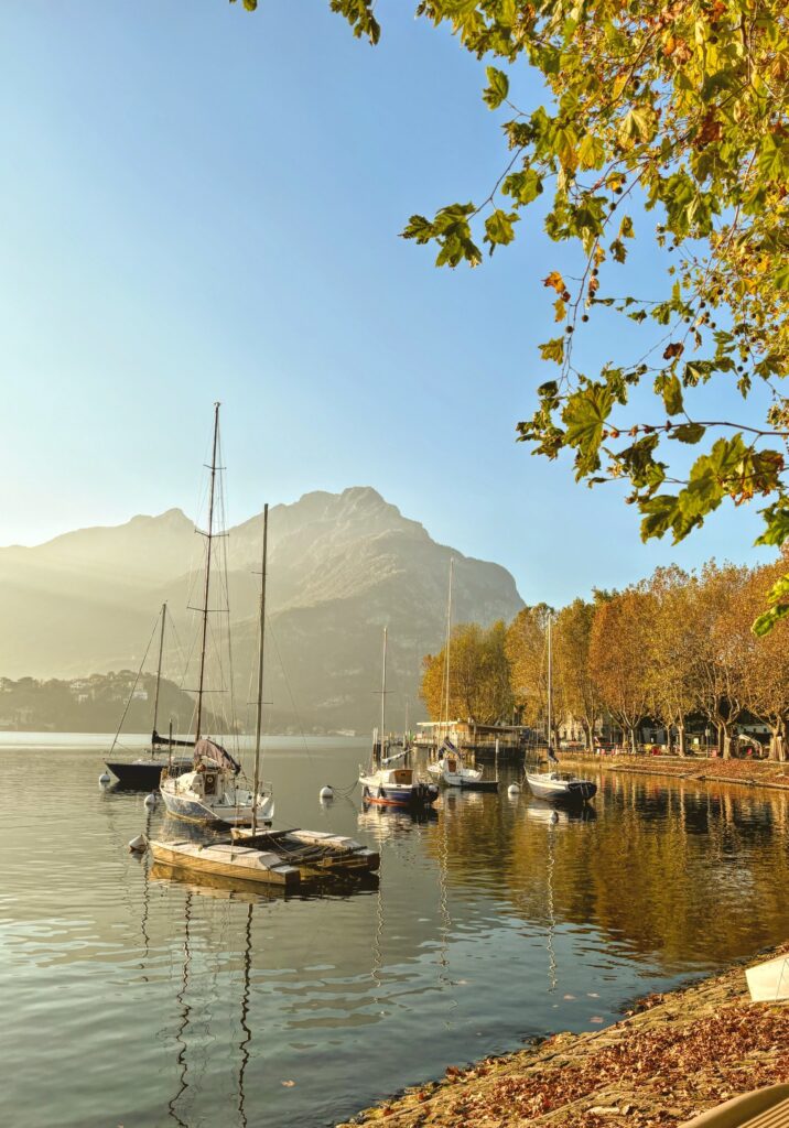 Sailboats gently floating on Lake Como at Lecco with golden autumn trees, calm reflections, and dramatic mountains in the background at golden hour.