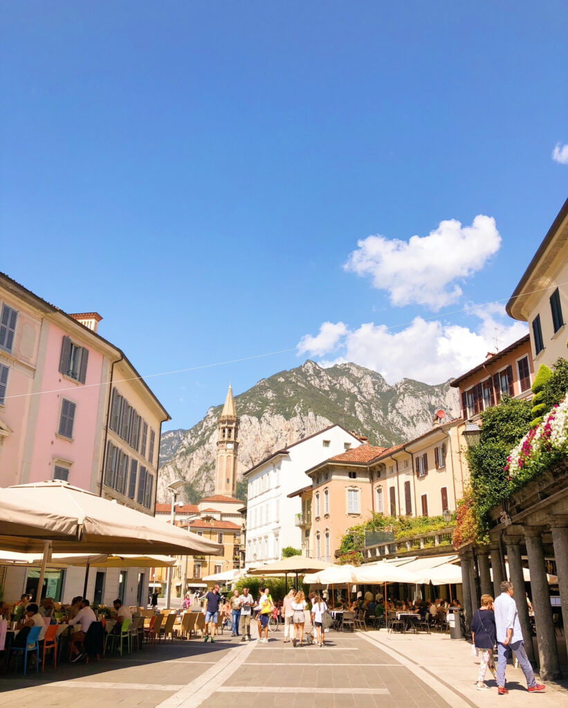 People dining and strolling through a sunlit piazza in Lecco, Northern Italy, with pastel buildings, a church tower, and mountain peaks in the background.