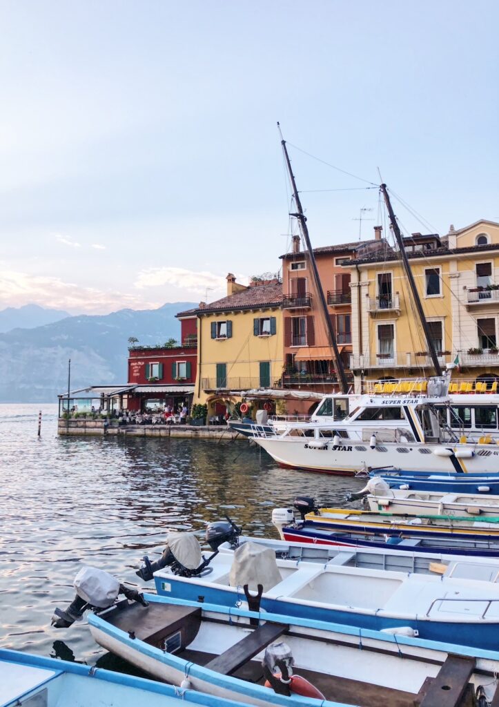 Colourful lakefront buildings and docked boats in Malcesine, Italy, with mountains in the distance and restaurants lining the peaceful harbour at sunset.