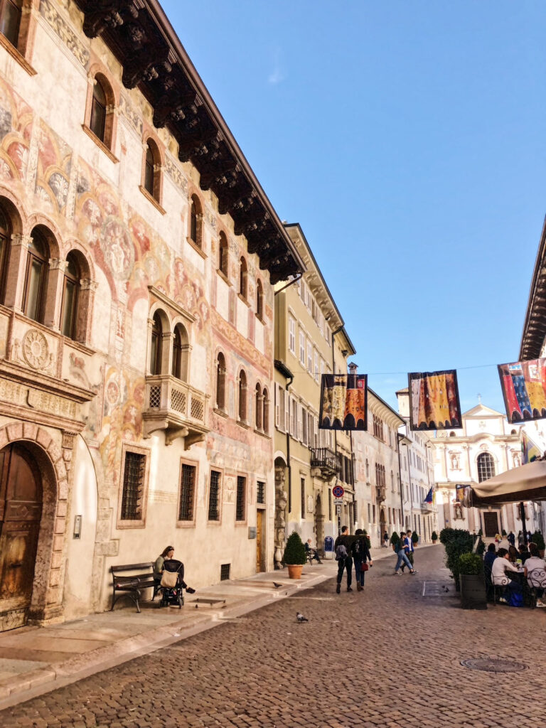 Ornate Renaissance-era frescoed buildings along a cobbled street in Trento, Northern Italy, with locals walking and dining under a clear blue sky.