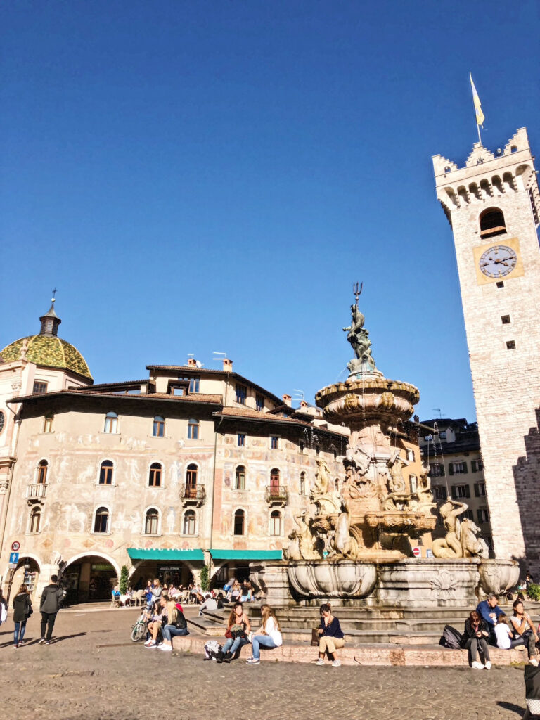 Neptune Fountain in Piazza Duomo, Trento, surrounded by frescoed palaces and the medieval Torre Civica, with people relaxing in the vibrant town square.