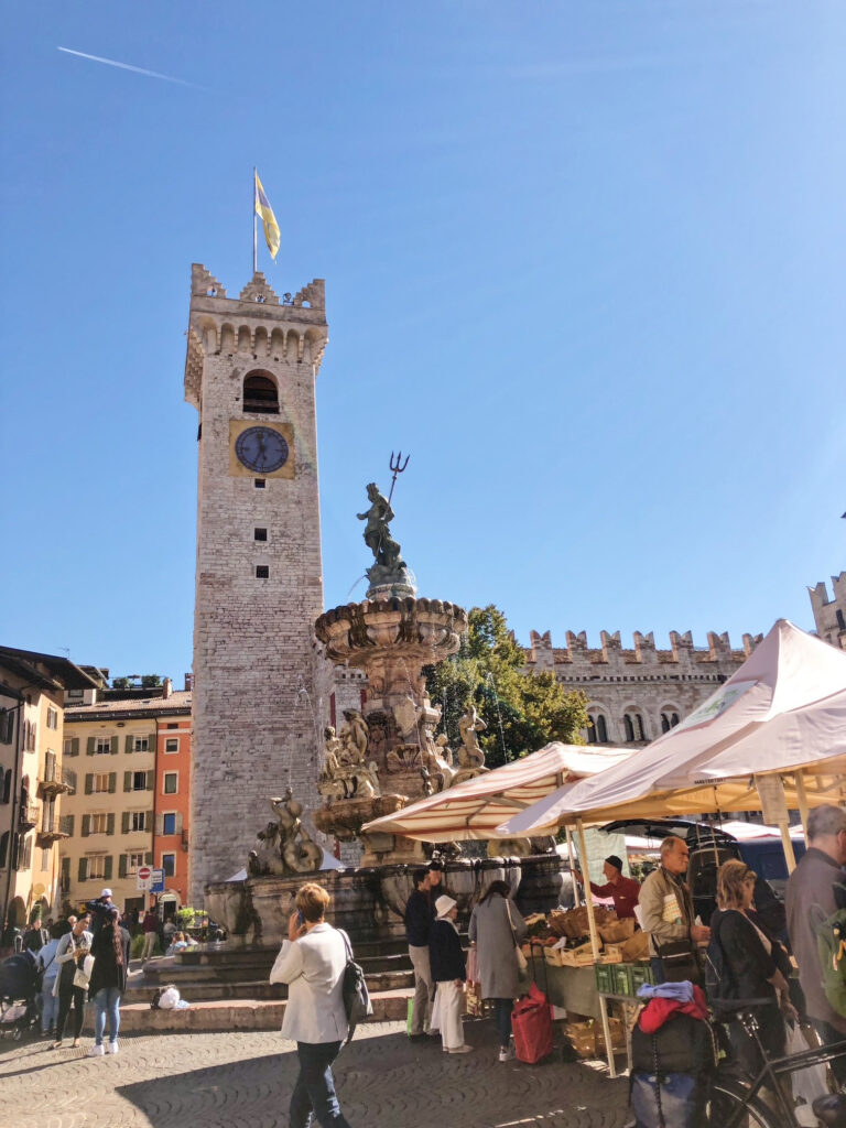 Lively farmers’ market in Trento’s Piazza Duomo beside the Neptune Fountain and Torre Civica, with stalls, shoppers, and historic architecture in the background.