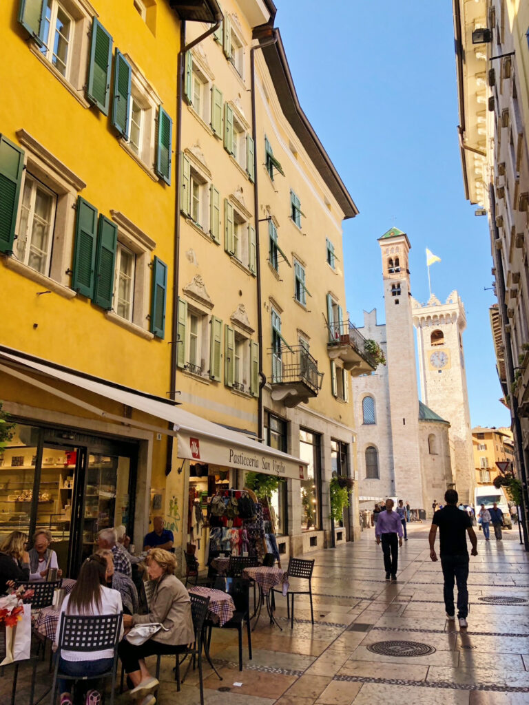 Colourful street view in Trento, Italy, with outdoor café tables, pastel buildings, and a view of Torre Civica rising at the end of the narrow walkway.