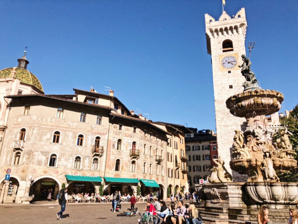 Frescoed buildings and the Neptune Fountain in Piazza Duomo, Trento, Italy, with Torre Civica and locals enjoying the lively square under a clear blue sky.